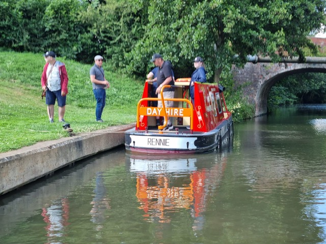 Canal boat with people on it, moored  at edge of canal. 